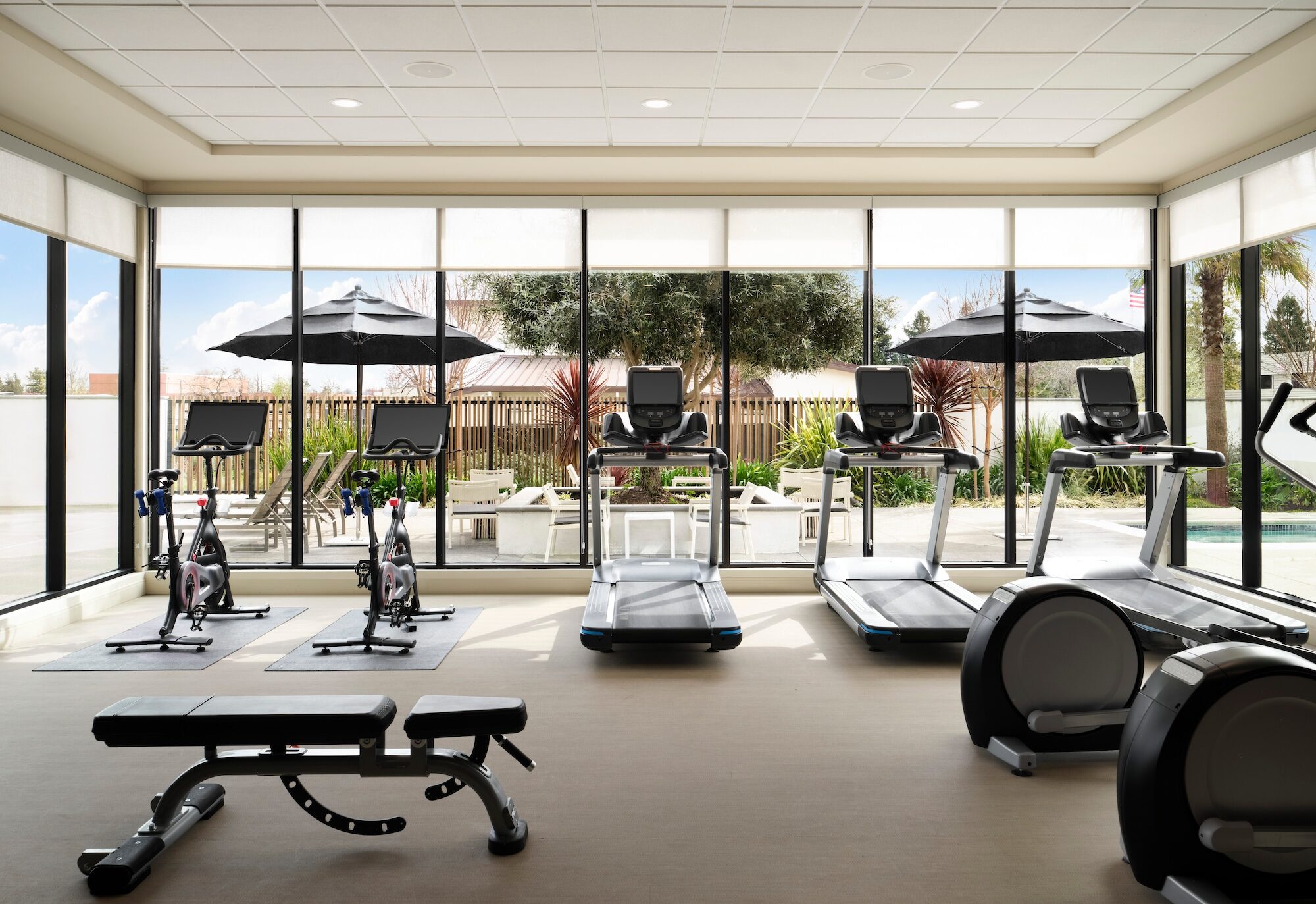 Hotel fitness center with treadmills, bikes, ellipticals, and a bench, facing floor-to-ceiling windows onto a pool patio