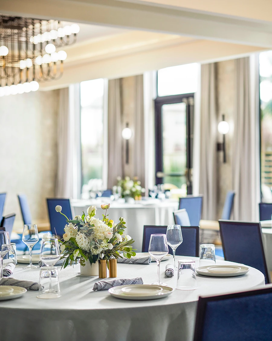 Banquet room set with round white-linen tables, blue chairs, white floral centerpieces, and tall windows with drapes