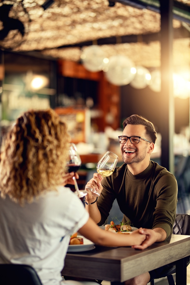 Smiling couple holding hands and toasting with red and white wine over dinner at a warmly lit restaurant table