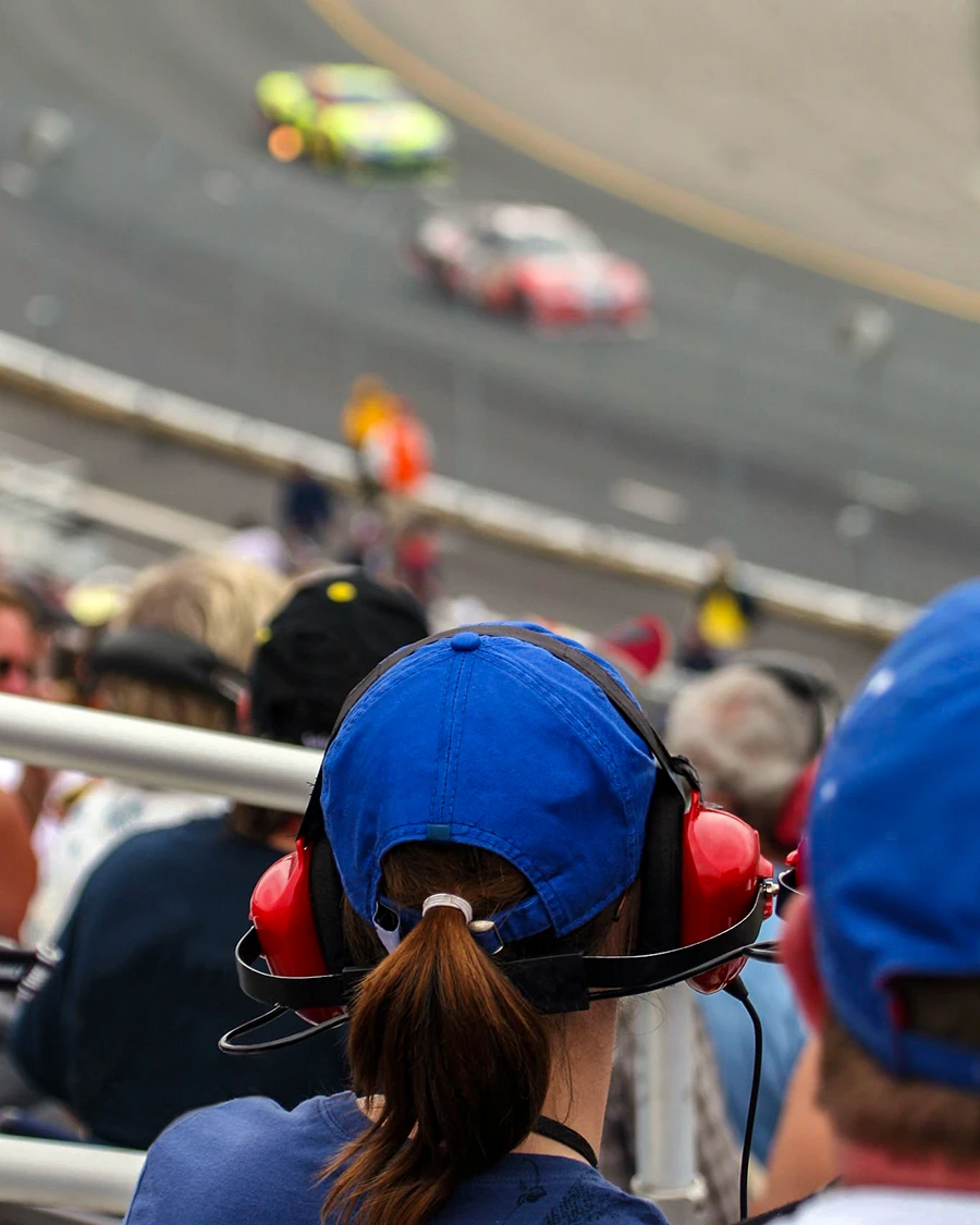 Spectator in a blue cap and red headset watches stock cars race past the grandstand at Sonoma Raceway