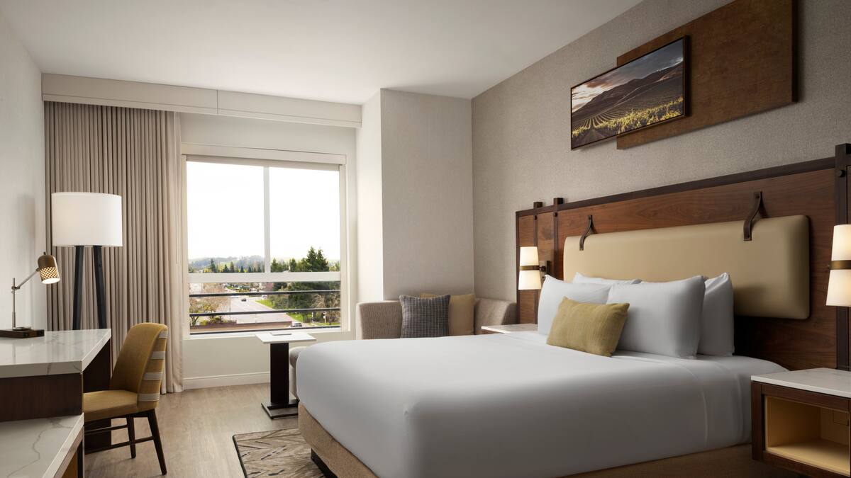 King hotel room with wood headboard, vineyard artwork, desk, sofa, and a large window overlooking trees and hills