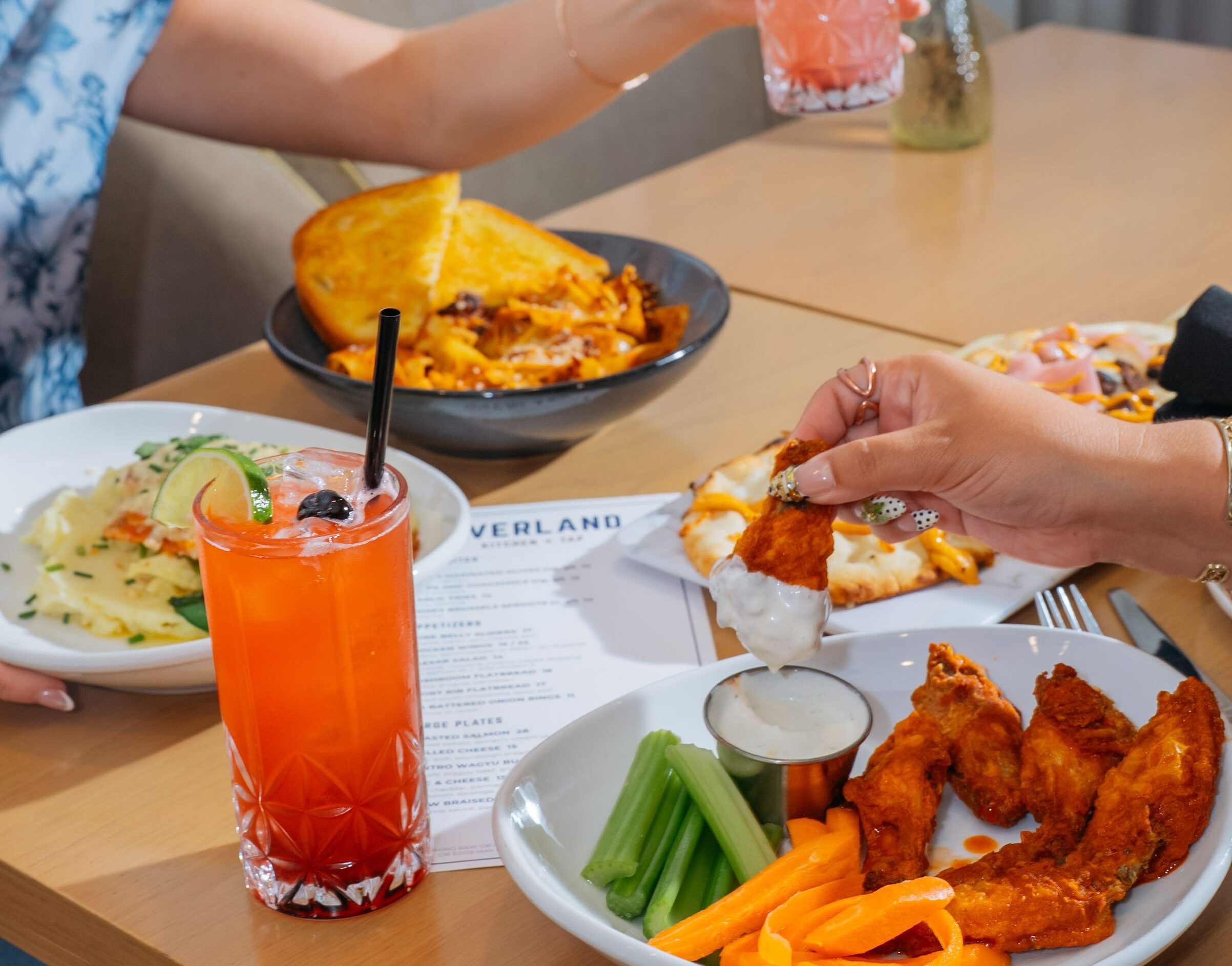 Diner dips a buffalo wing in ranch beside a red cocktail, mac and cheese, and flatbread on an Overland Kitchen + Tap menu